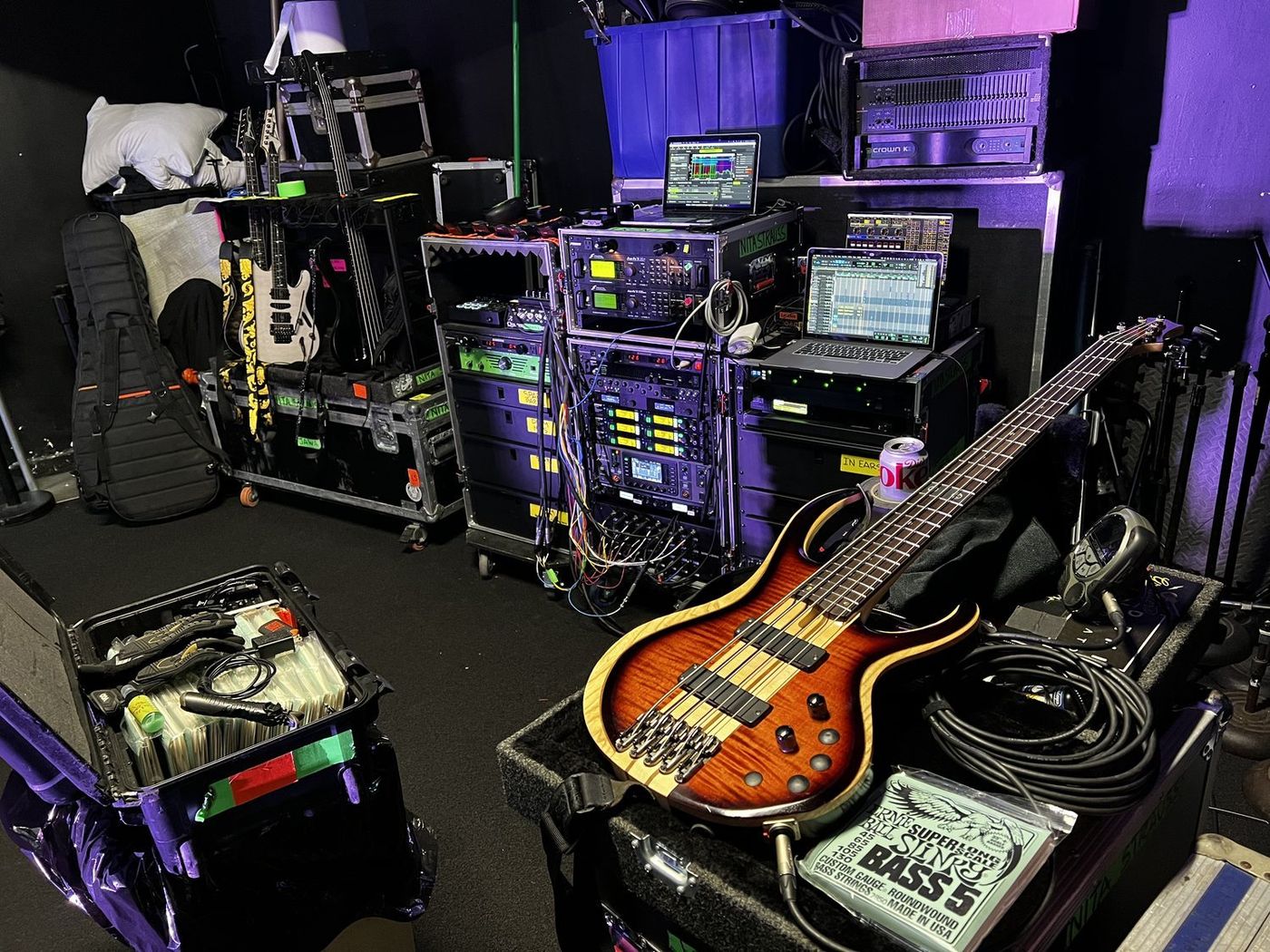 Side stage: rack cases, two laptops running the rigs, guitar cases, and an amber Ibanez BTB five-string laid on a case beside a pack of Ernie Ball Super Slinky Bass 5 strings